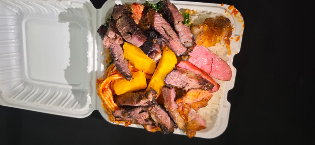 Takeout box packed with sliced churrasco, sausage, chicken, fried cassava, rice, pasta and collard greens