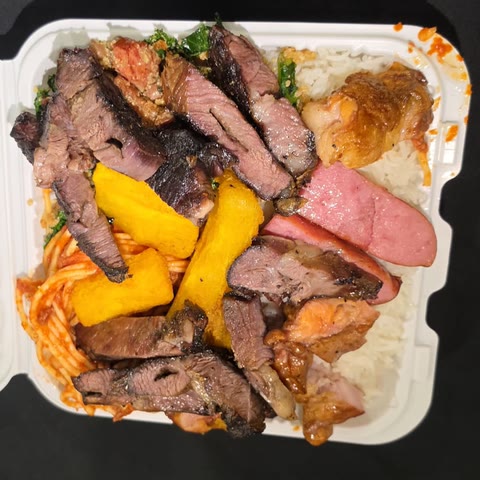 Takeout box packed with sliced churrasco, sausage, chicken, fried cassava, rice, pasta and collard greens
